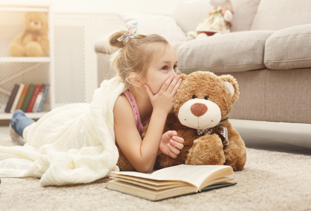 A young girl lying on the floor with a book in front of her whispers to her teddy bear.