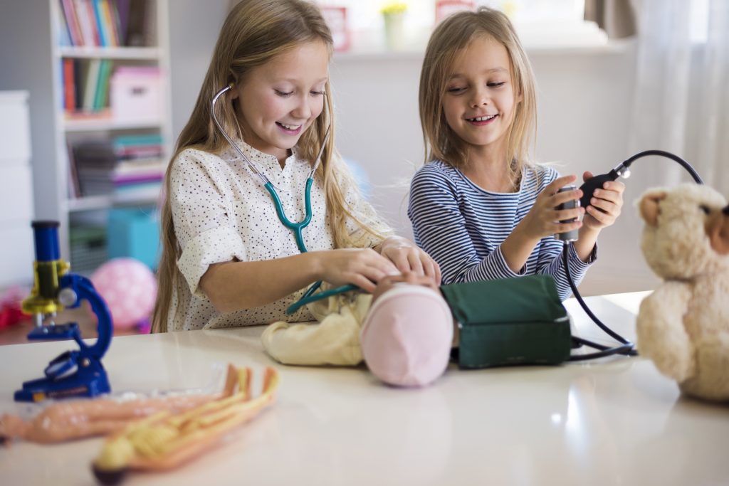 A young girl uses a stethoscope on a doll, while another young girl looks on, holding a blood pressure cuff.