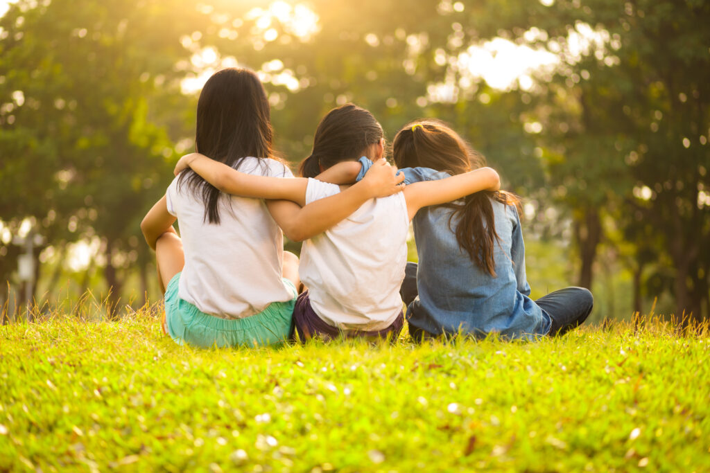 Three young girls sitting on the grass, arms around each other's shoulders, facing away from the camera