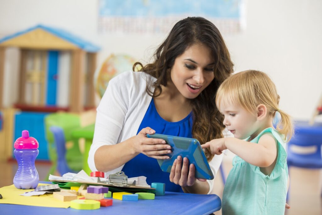 A young child touches a tablet held by a childcare educator