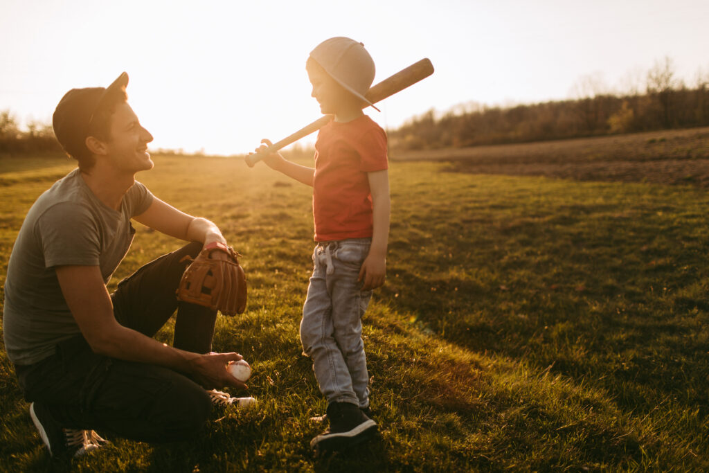A father with a baseball and glove and his son with a bat over his shoulder smiling at each 