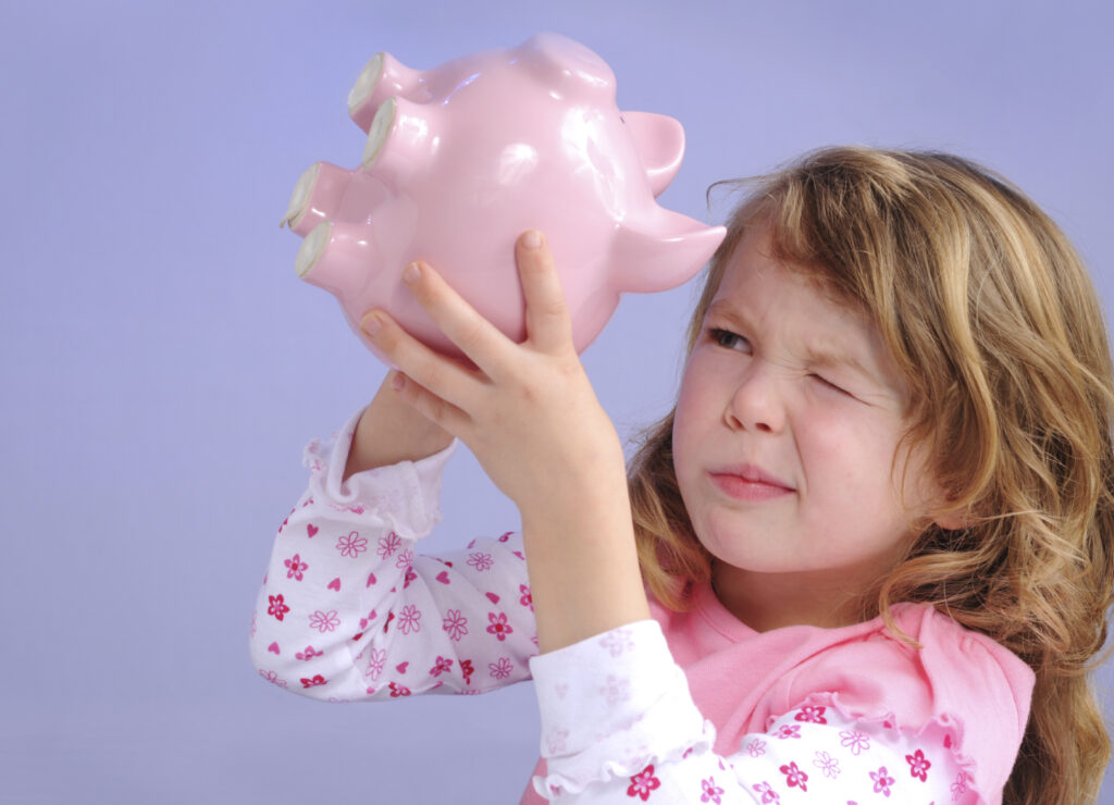 A young girl holding a piggy bank upside, down, squinting into the slot with one eye closed.