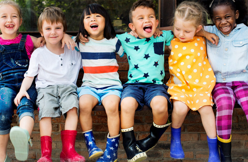 Six young children of various ethnicities, sitting a la line with their arms around each other, smiling