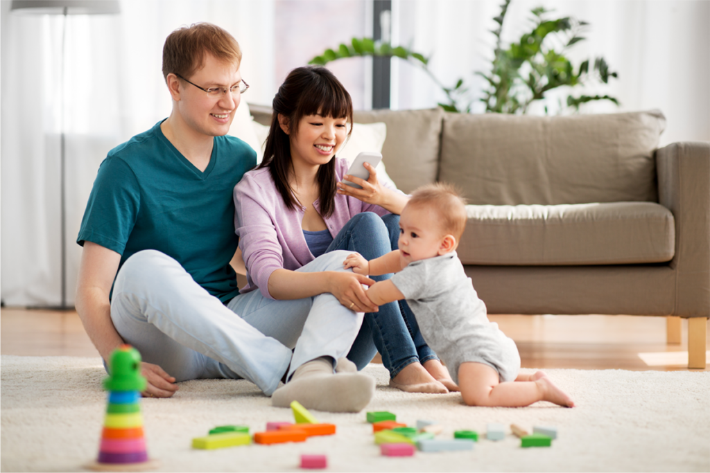 A mother and father sit on the floor while their baby attempts to stand while holding on to their legs