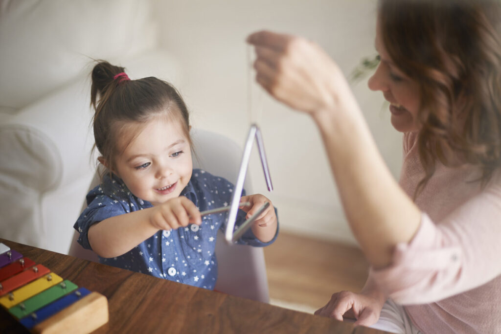 A mother holds a triangle while her daughter strikes it while a xylophone sits unused on the table, indicating the mother is helping promote agency in early childhood