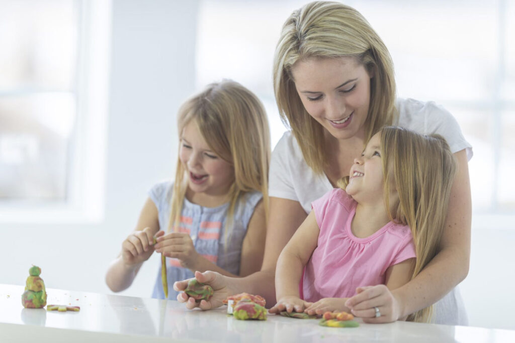 A mother smiling at her daughter sitting in her lap playing with playdough with her sister. Perhaps the mother has just given the children proper praise for their efforts