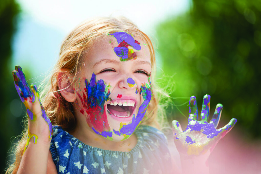 A very happy young girl, with her hands and face covered in different coloured paint, another example of unstructured play