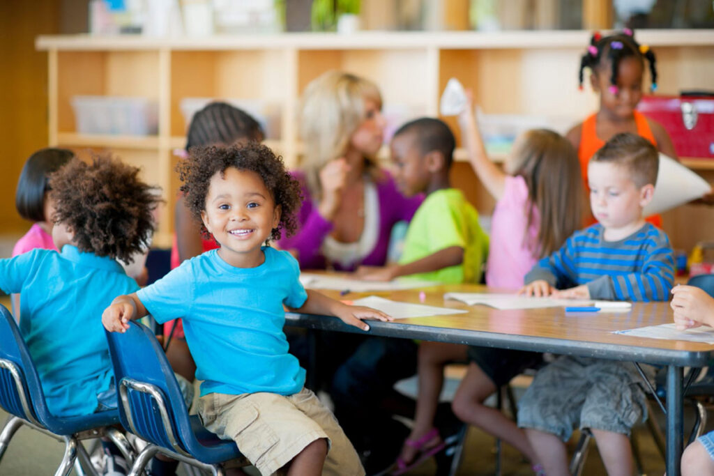 A young boy in a classroom looking at the camera, with the other students and teacher out of focus in the background