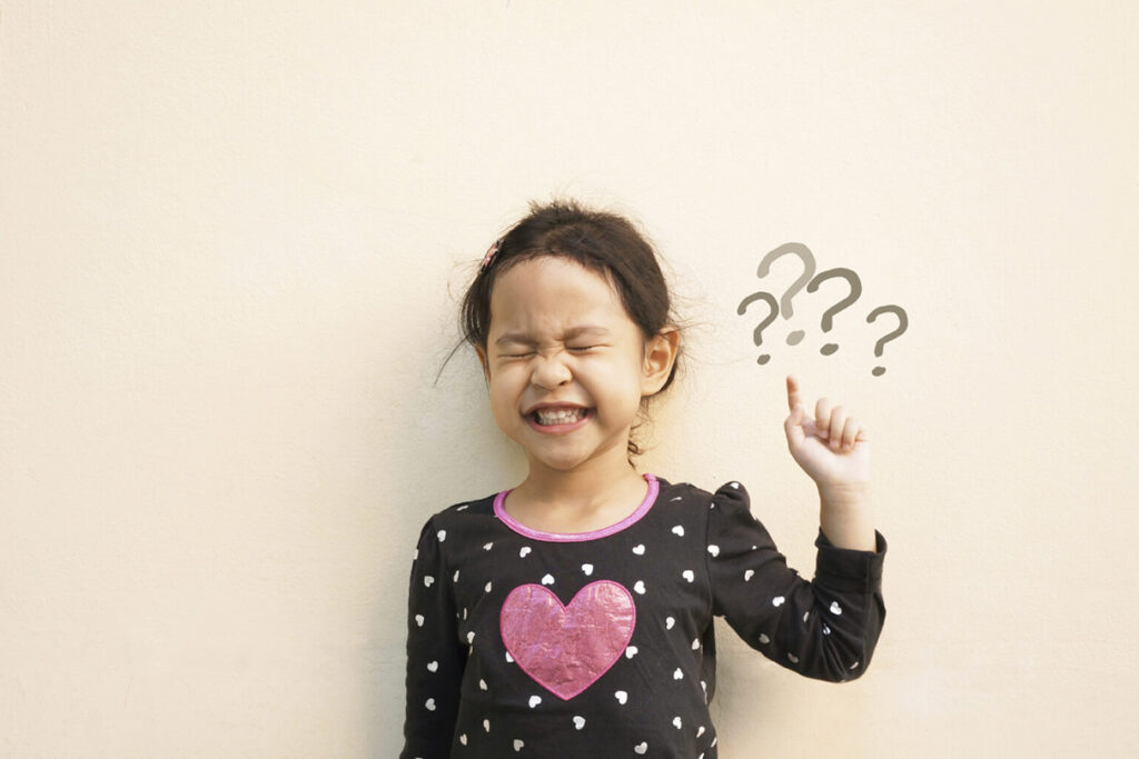 A young girl, eyes scrunched shut, pointing over her left shoulder at question marks drawn on the wall.
