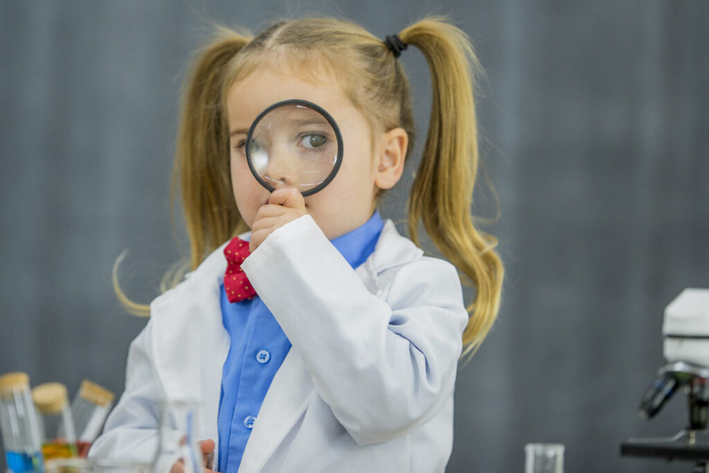 A young girl, dressed in a lab coat, staring at the camera through a magnifying glass.