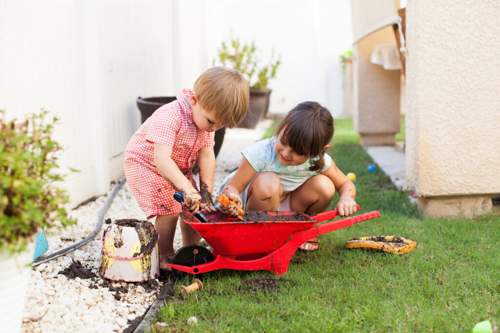 Two young children playing with mud in a small red wheelbarrow, representing the need to remind children to wash their hands, a good hygiene practice