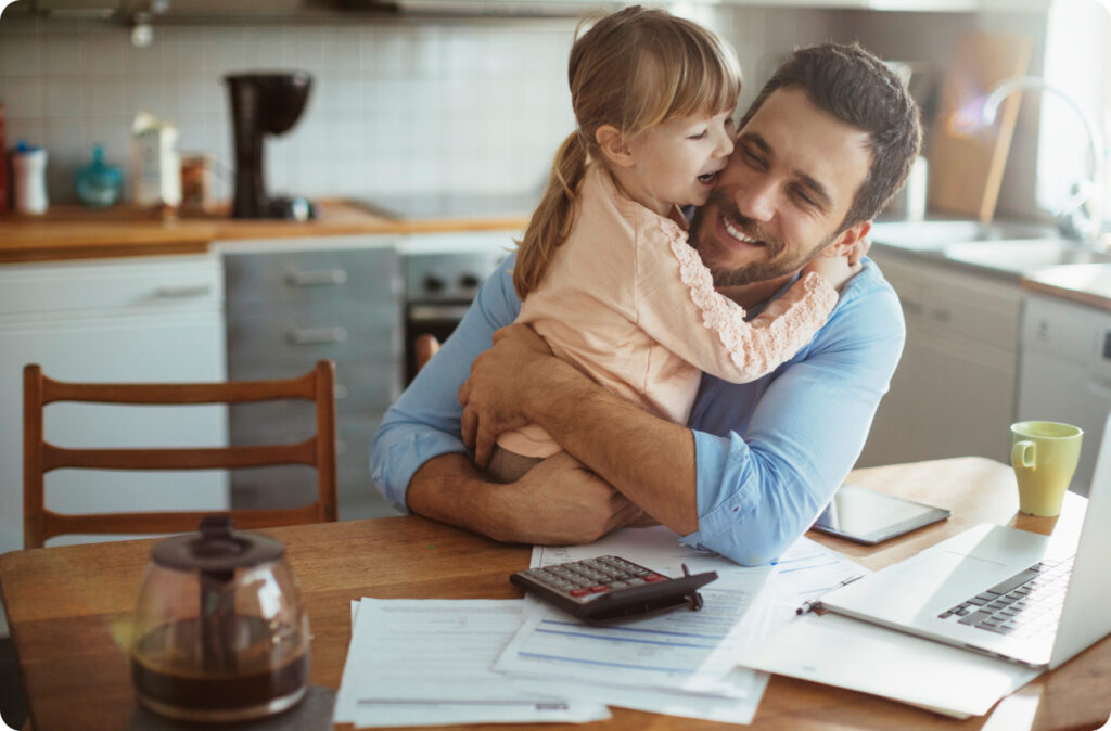 A father hugs his young daughter as he sits at a kitchen table, strewn with papers, a calculator and a laptop computer.