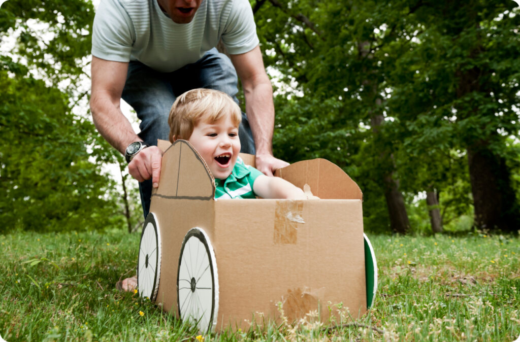 Father playing with his son riding in a recycled cardboard toy car, a great way to encourage sustainability in childcare