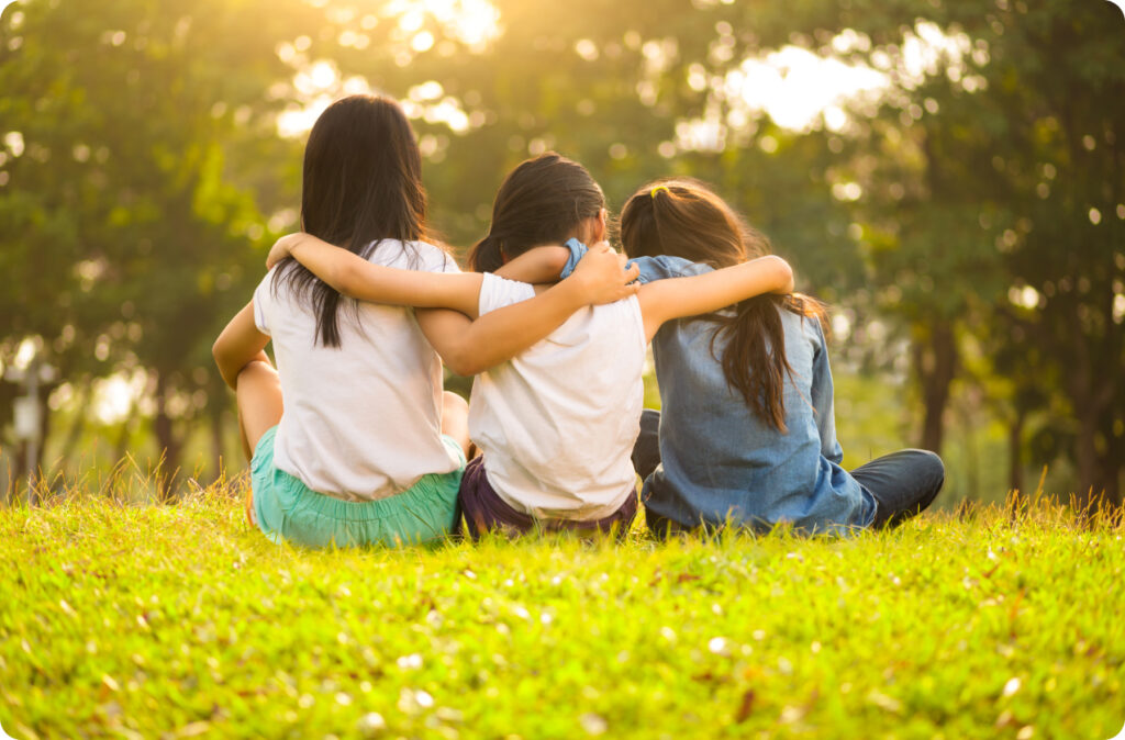 Three young girls, facing away, sitting on the grass with their arms around each other.