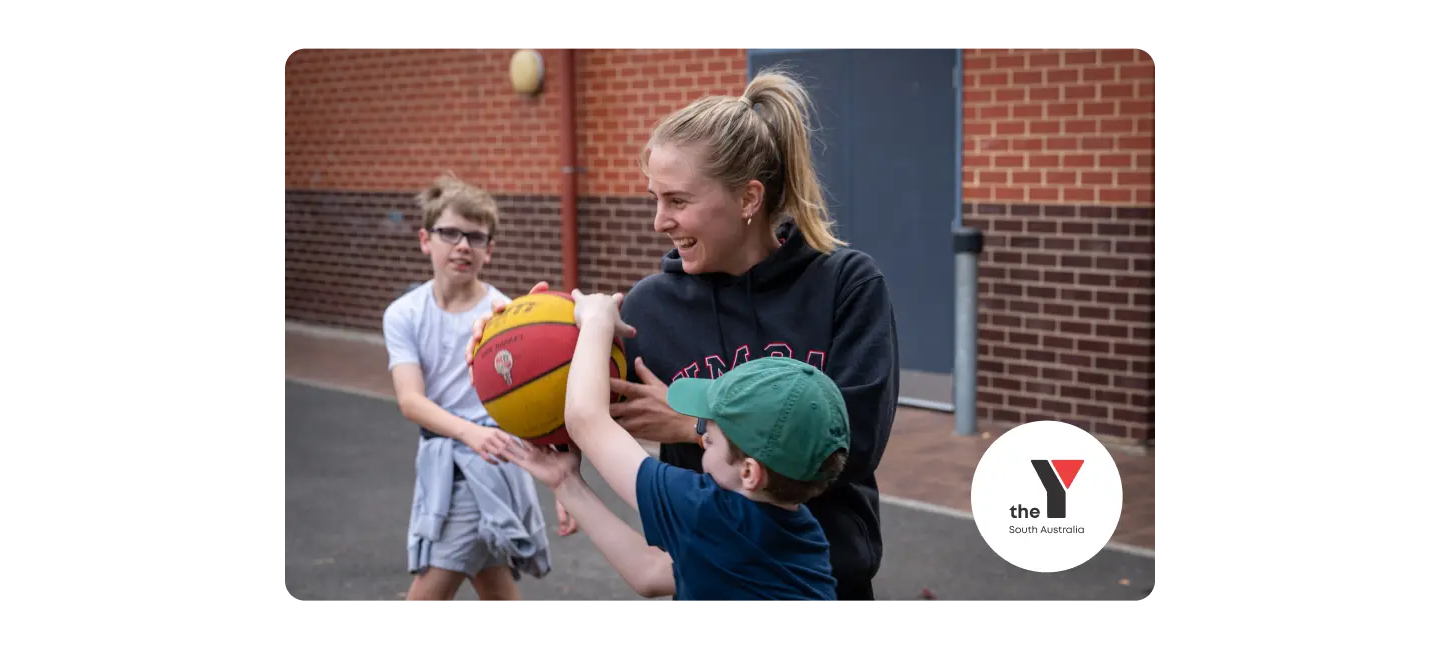 Educator playing basketball with two children with the YMCA South Australia logo in the bottom corner