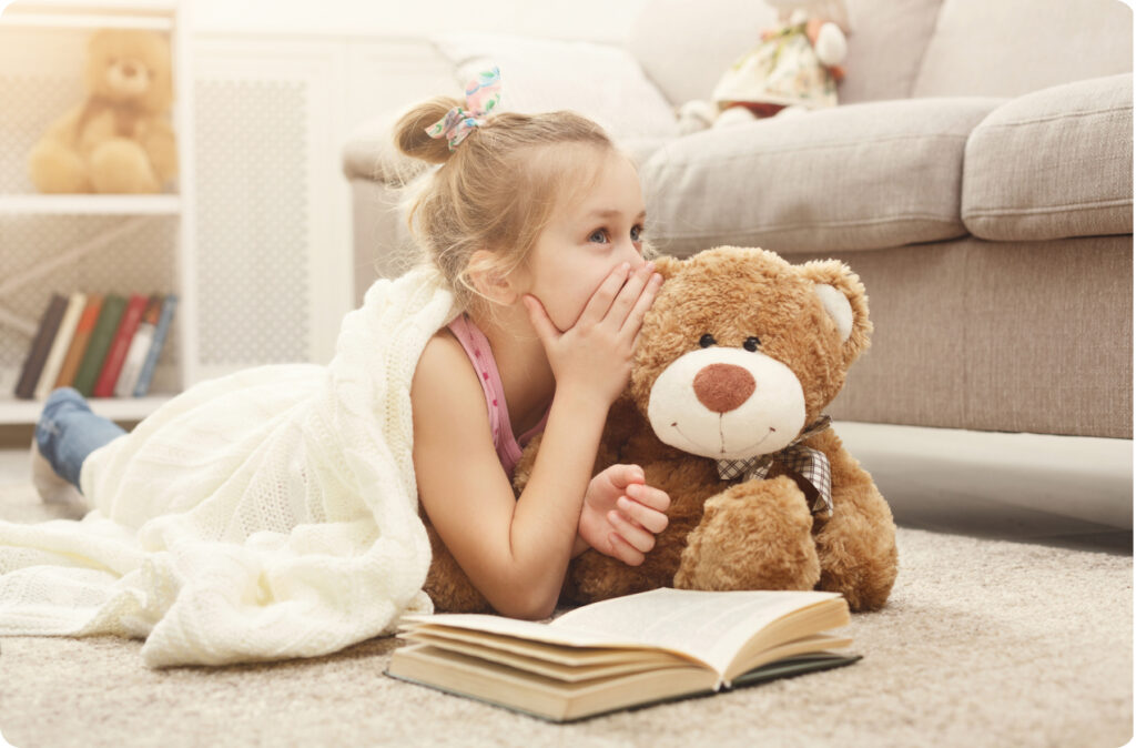 A young girl lying on the floor with a book in front of her whispers to her teddy bear, perhaps about a children's book by an Australian author