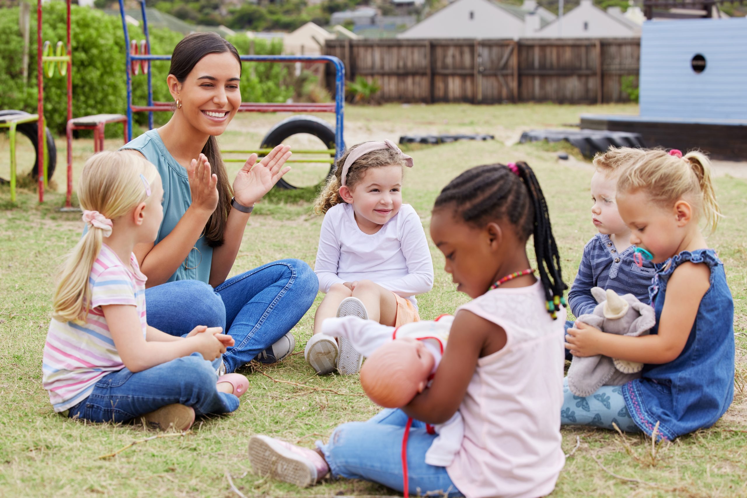 An educator and children sitting on some grass. The educator is clapping as a young girl cradles a doll.