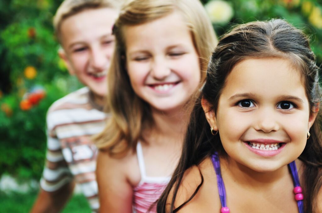 Young girl smiling at the camera with two children in a blurred background also smiling