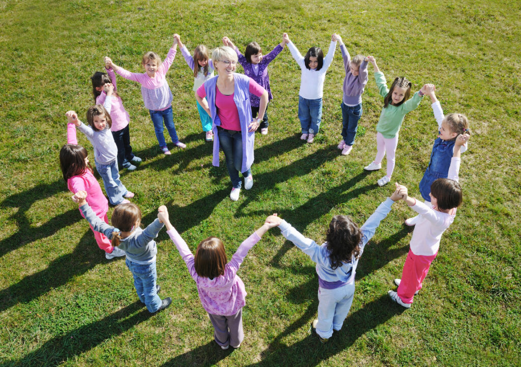 An educator surrounded by children in a grass field, representing a supportive community, one of many strategies for preventing teacher burnout in early childhood education