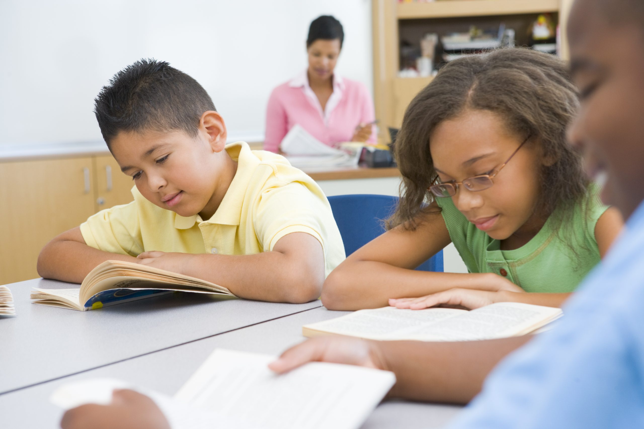 Children read at their desks as the teacher looks on in the background