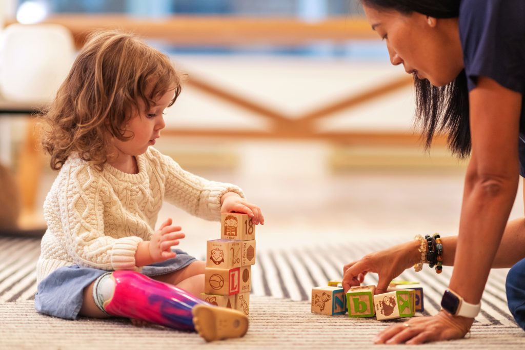 A young girl with a prosthetic leg stacks blocks while an educator looks on