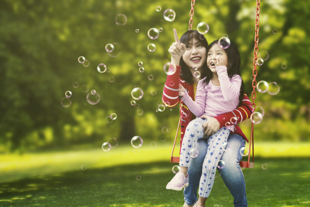 A mother and daughter on a swing, smiling and pointing at multiple bubbles in the air.
