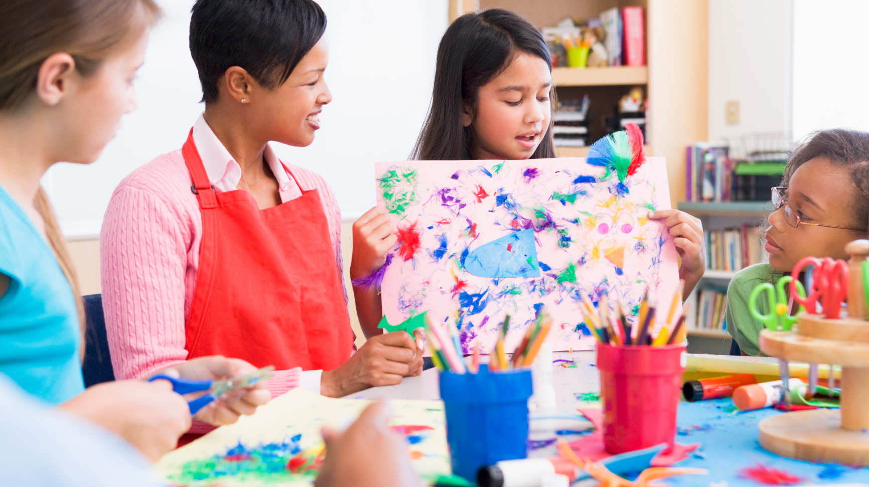 An educator and fellow students look on as a young girl holds up an abstract painting