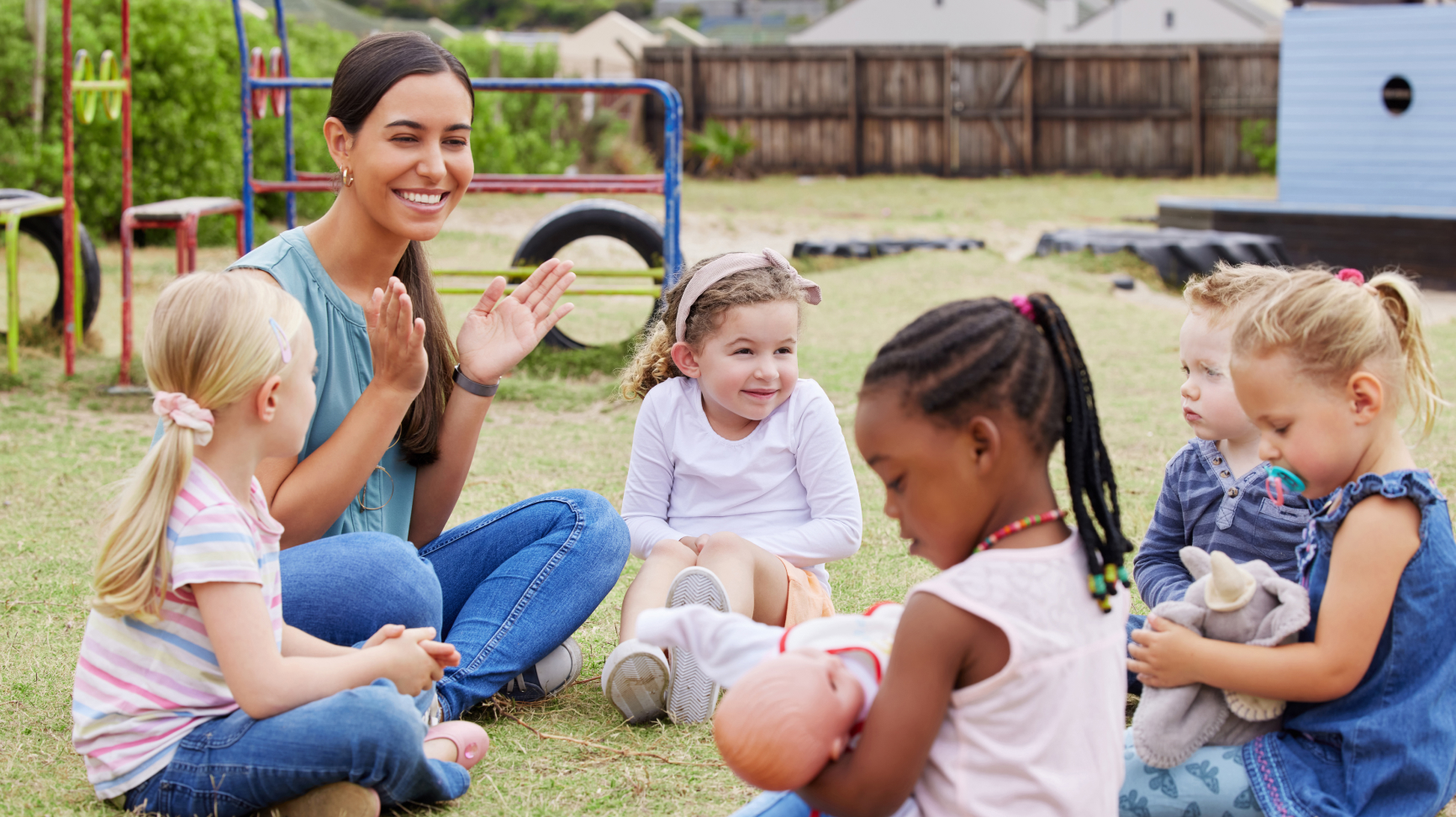 Children sit on the the grass, some holding dolls as an educator smiles and claps