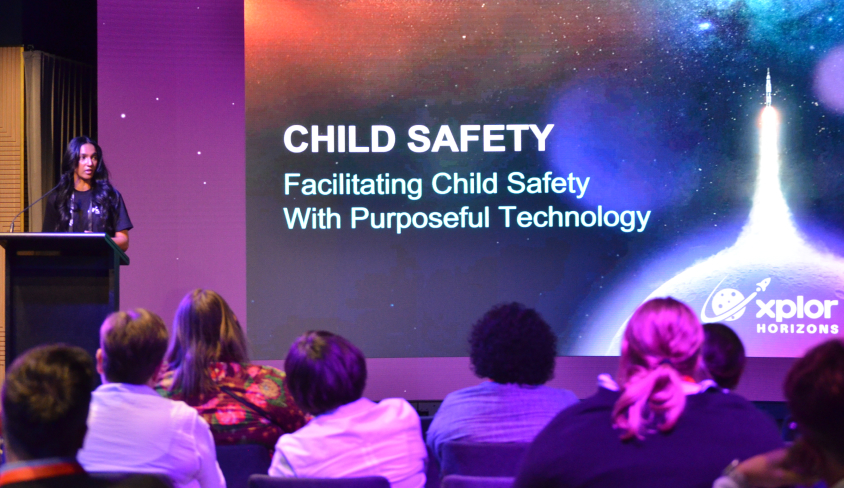 Presenter at the Xplor Horizons event speaking about child safety, with a slide in the background saying "Facilitating Child Safety with purposeful technology"